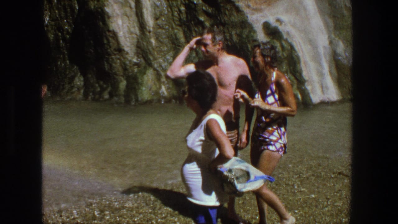 Adults in swimsuits and summer clothing stand by shallow water below a rocky waterfall.