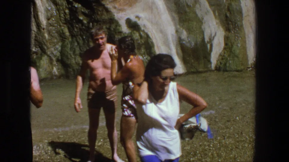 A woman in sunglasses passes near the camera while friends remain by the waterfall in the 1969 Utah home movie.