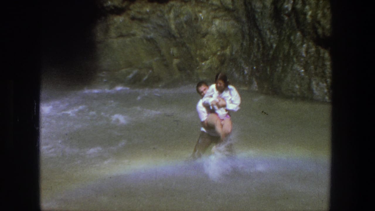 In 1969 Utah home-movie footage, a man in a white shirt carries a woman through a shallow canyon stream.