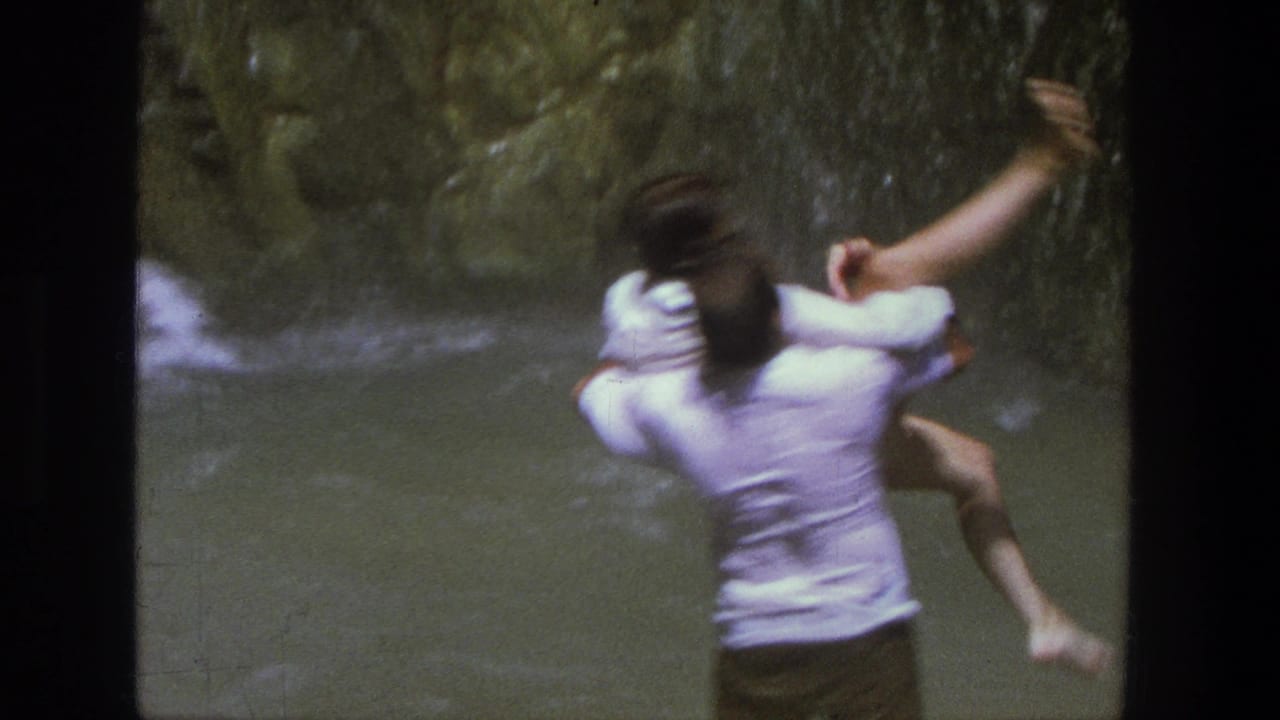Man carries a woman in rushing water with canyon vegetation behind them.