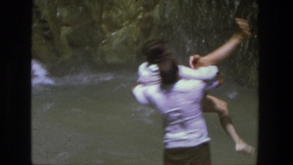 A man carries a woman through a Utah waterfall stream, seen from behind in soft 8mm color footage.