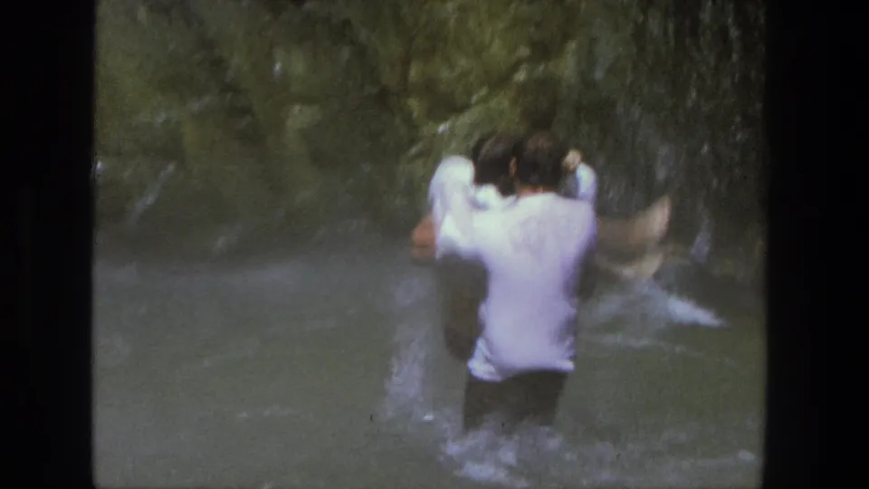 A man in a white shirt carries a woman through splashing water in a 1969 Utah canyon home movie.