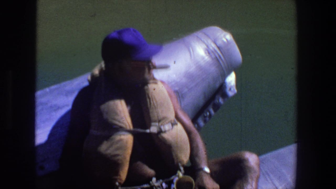 Man in a purple cap and tan life jacket seated on a boat beside green water.