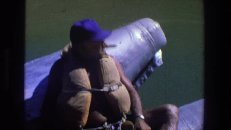 Adult man in sunglasses, purple cap, and tan life jacket sits on a boat during a 1969 Utah water outing.