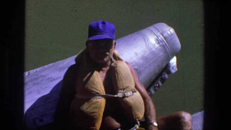 Adult man faces the camera from a boat, wearing sunglasses, a purple cap, and a tan life jacket in 1969 Utah footage.