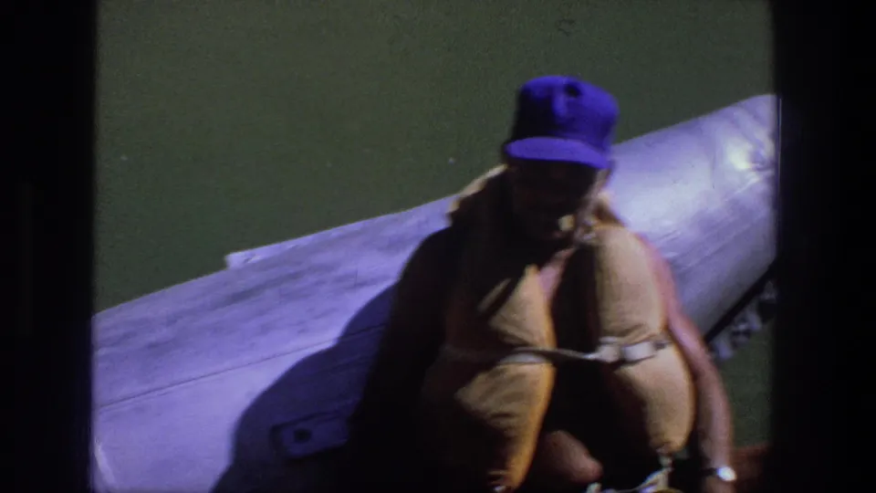 Adult man in a tan life jacket and purple cap remains seated on the boat, with green water and tubing behind him.