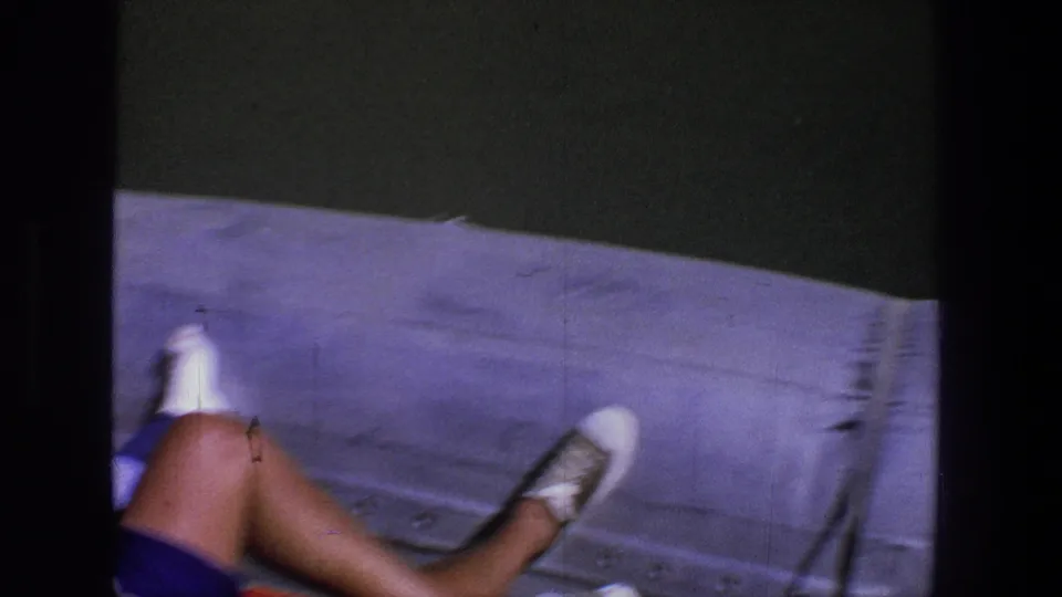 A person reclines on the boat with legs and white shoes visible, while green water fills the background.