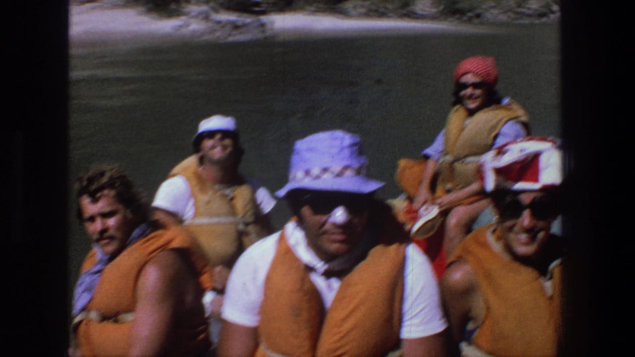 Adults in orange life vests and hats sit on a river raft in bright sun.