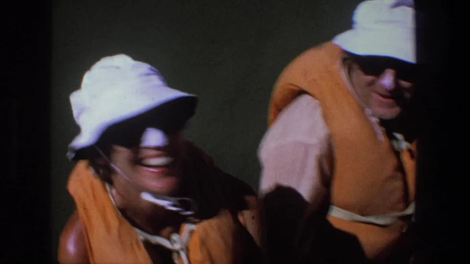 Close handheld frame from a 1969 Utah river trip showing adults in orange life vests and sun hats near dark water.