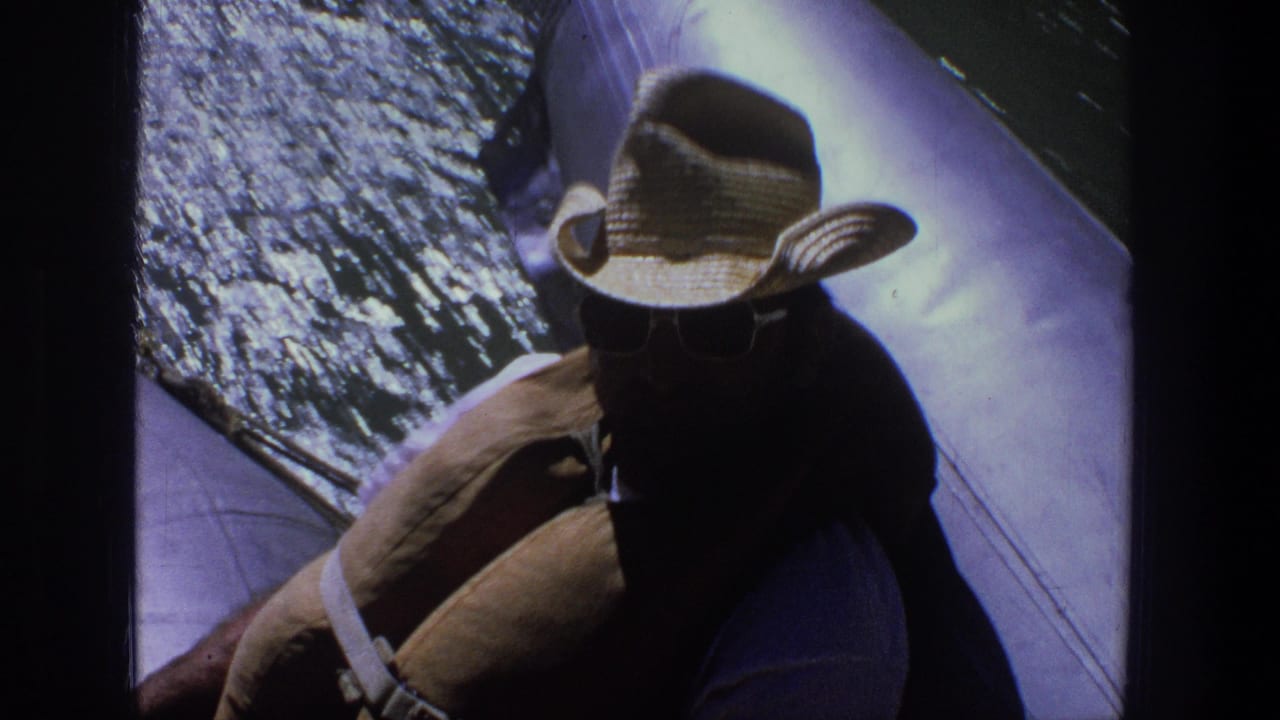 Man in a straw cowboy hat and sunglasses seated in a boat beside sparkling water.