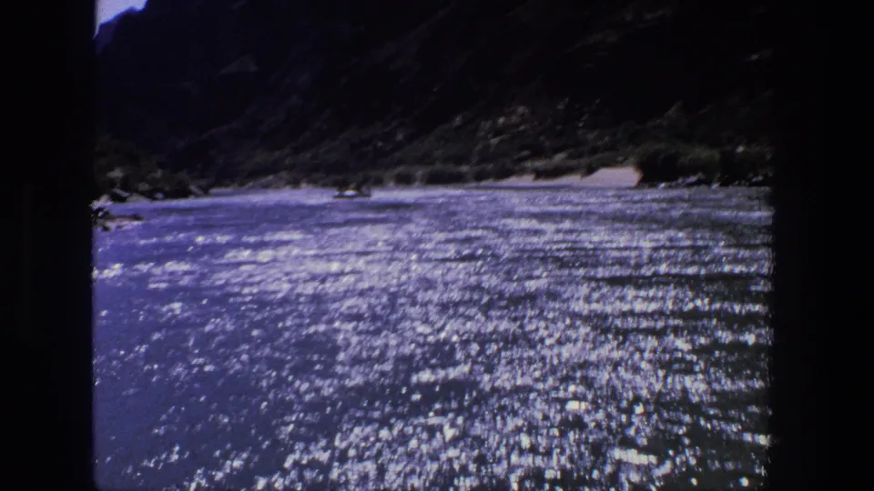 1969 Utah 8mm frame with shimmering river water in the foreground and dark canyon slopes rising behind a light sandy shoreline.