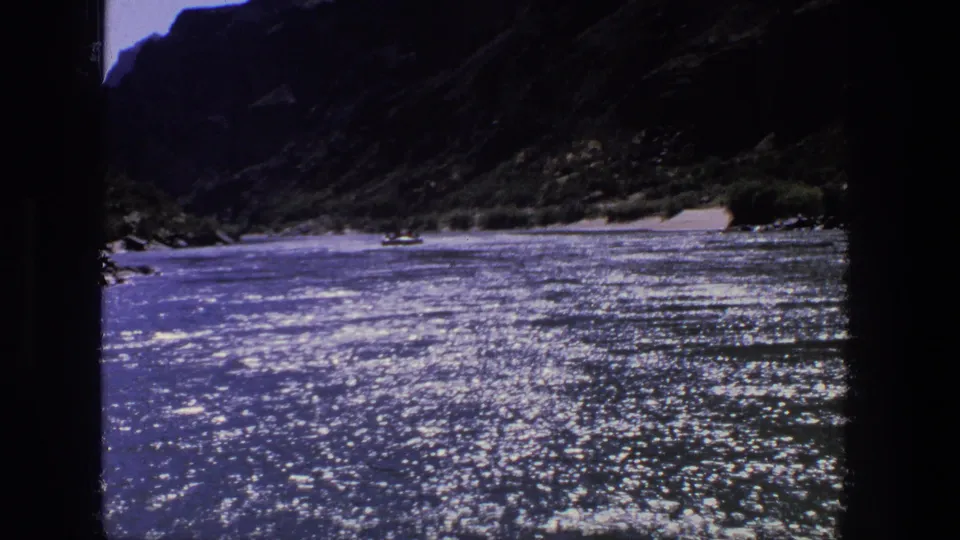 1969 Utah 8mm frame of a sparkling canyon river with a distant boat, steep dark walls, sandy banks, and sparse green vegetation.