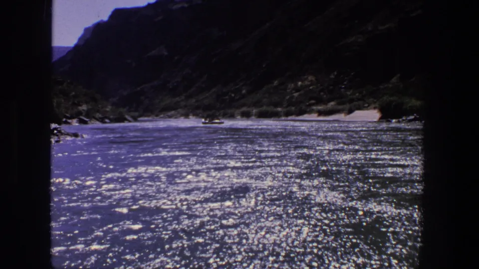 Sunlit 1969 Utah river canyon scene on 8mm film, showing shimmering water, shadowed cliffs, and a small boat far away.