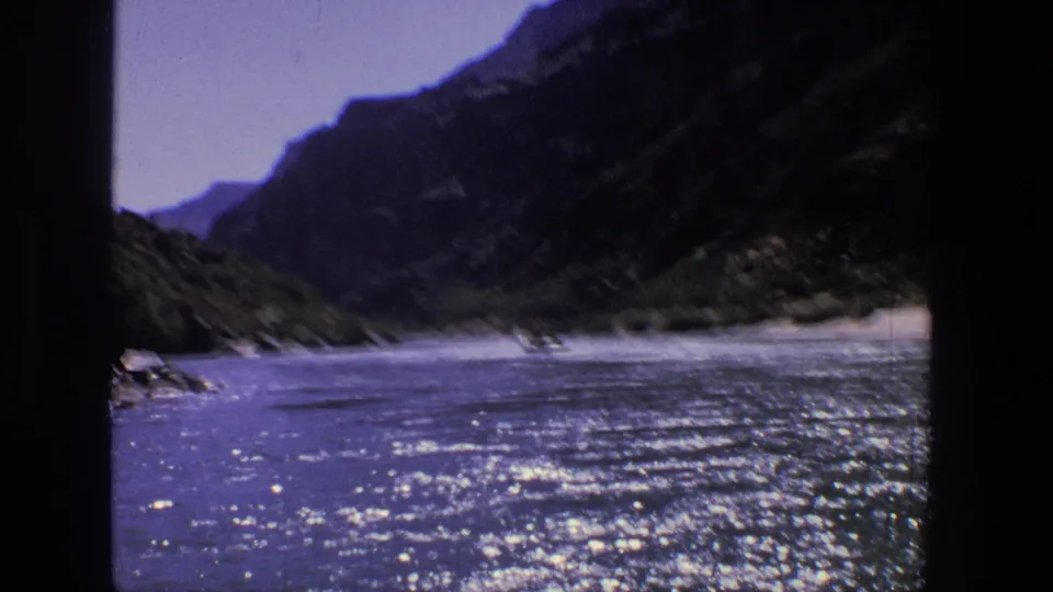 Soft handheld 8mm frame from 1969 Utah showing a bright river surface, sandy bank, and steep canyon walls under clear sky.