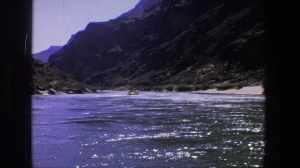 1969 Utah 8mm frame of sunlit river ripples, rocky canyon slopes, green banks, and a small boat in the distance.
