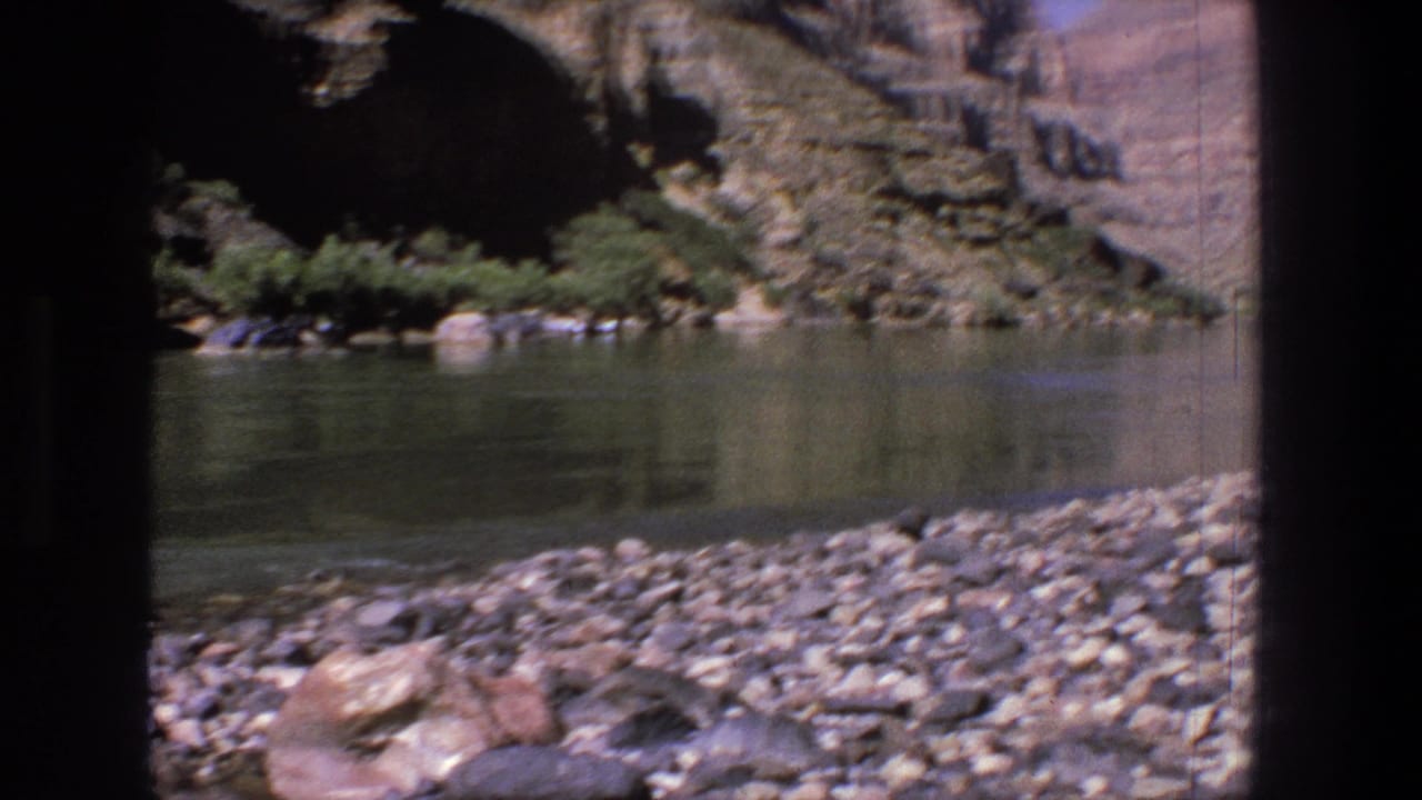 Rocky riverbank beside green water below shaded canyon walls.