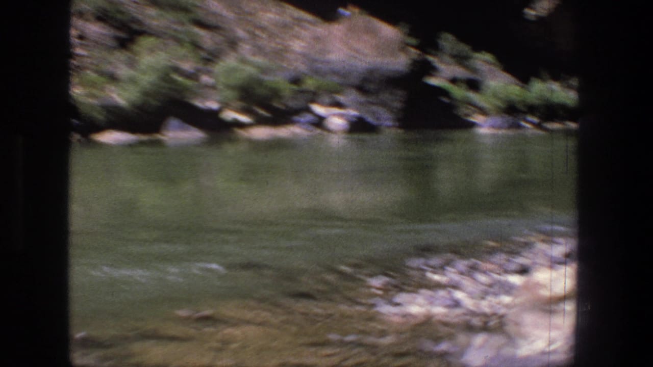 1969 Utah canyon river view with green water, scattered rocks, and low vegetation along the far bank.