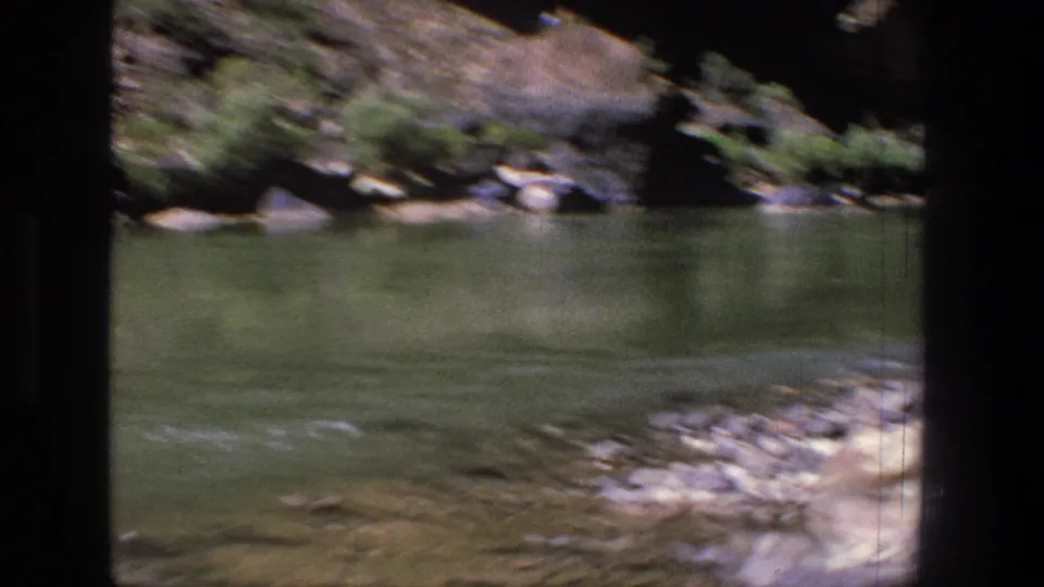 1969 Utah canyon river view with green water, scattered rocks, and low vegetation along the far bank.