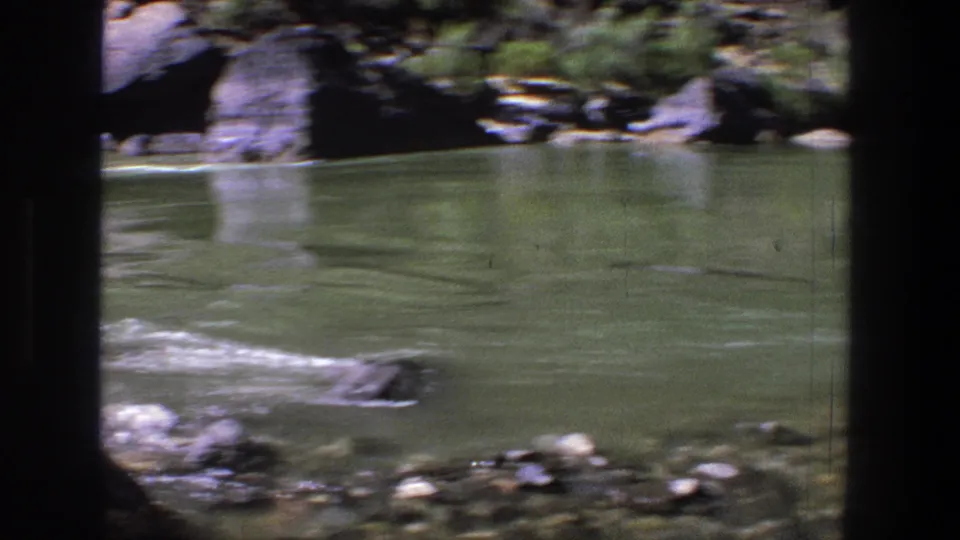 Vintage 8mm Utah river frame showing calm green water, a stony bank, and shadowed canyon rock in the background.