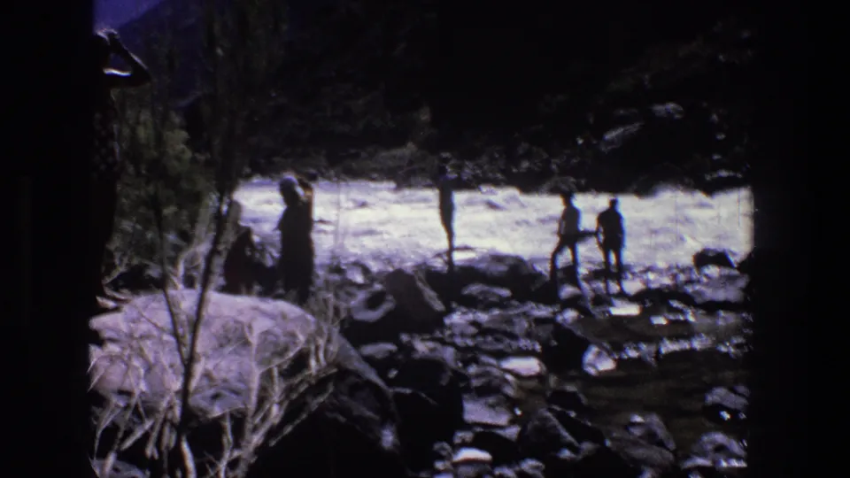 1969 Utah home-movie frame of several distant people on a rocky riverbank, watching bright whitewater in deep canyon shadow.