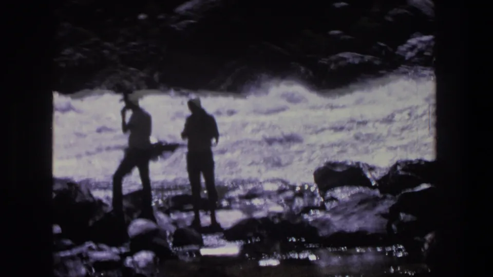 1969 Utah 8mm frame showing two dark figures on a rocky riverbank against bright moving water.