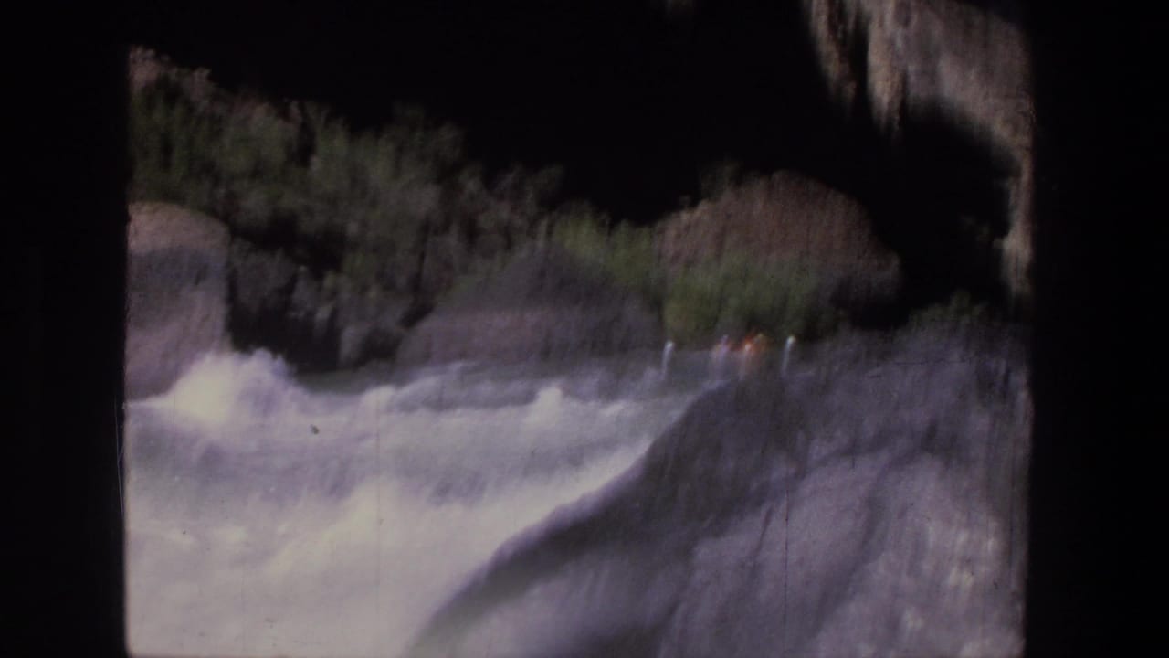 Small raft with orange life jackets moving through whitewater beside dark rocks.