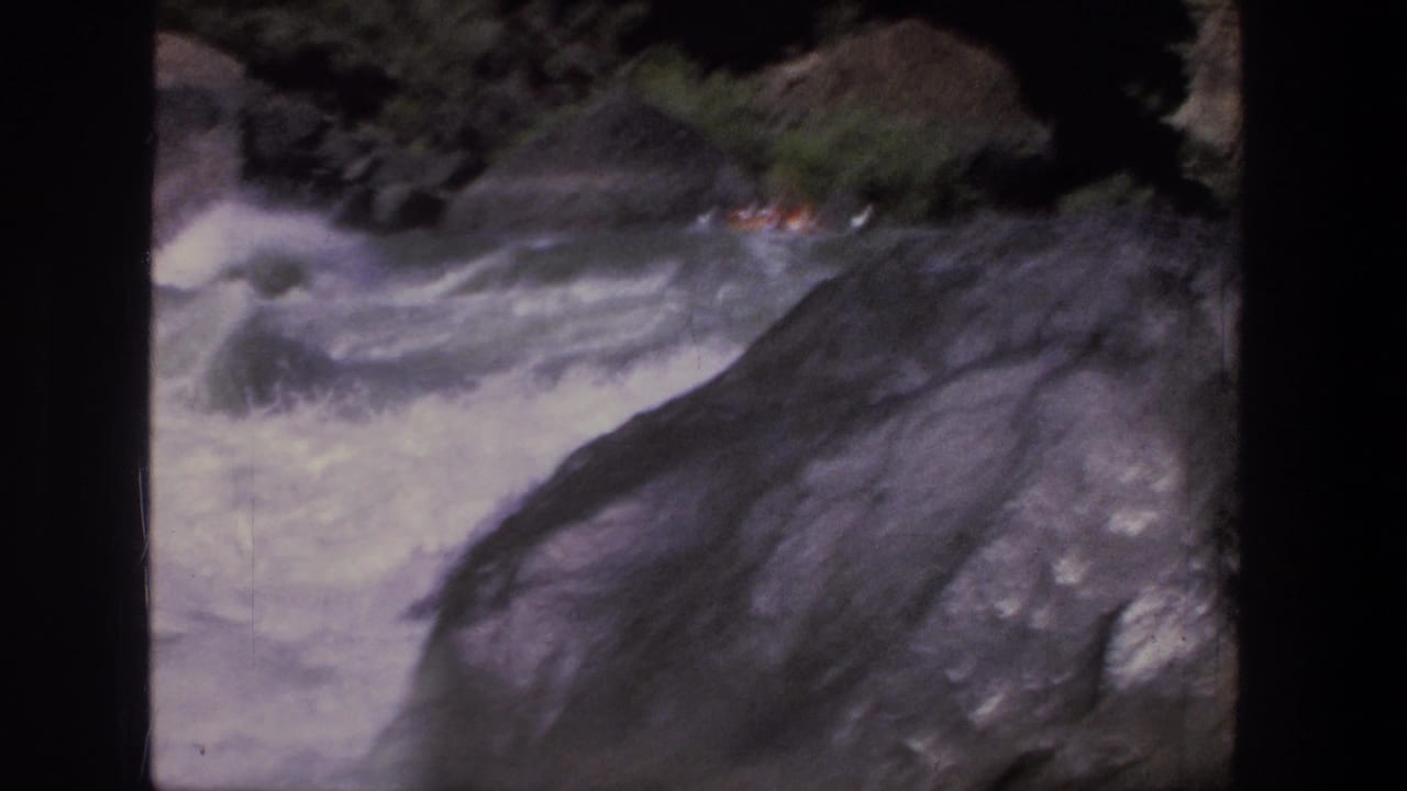 A distant raft with riders in orange life jackets moves through Utah rapids, partly obscured by a large dark rock.