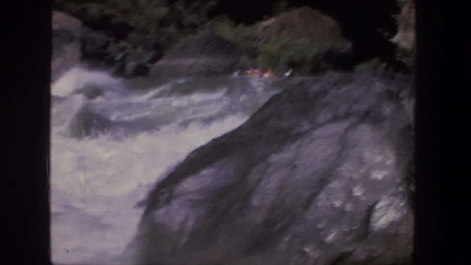 A distant raft with riders in orange life jackets moves through Utah rapids, partly obscured by a large dark rock.