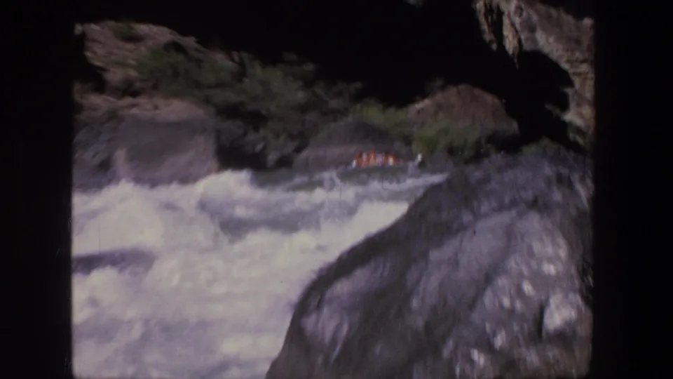 A small raft with orange-clad riders continues through Utah whitewater, framed by wet rocks, shadows, and low vegetation.