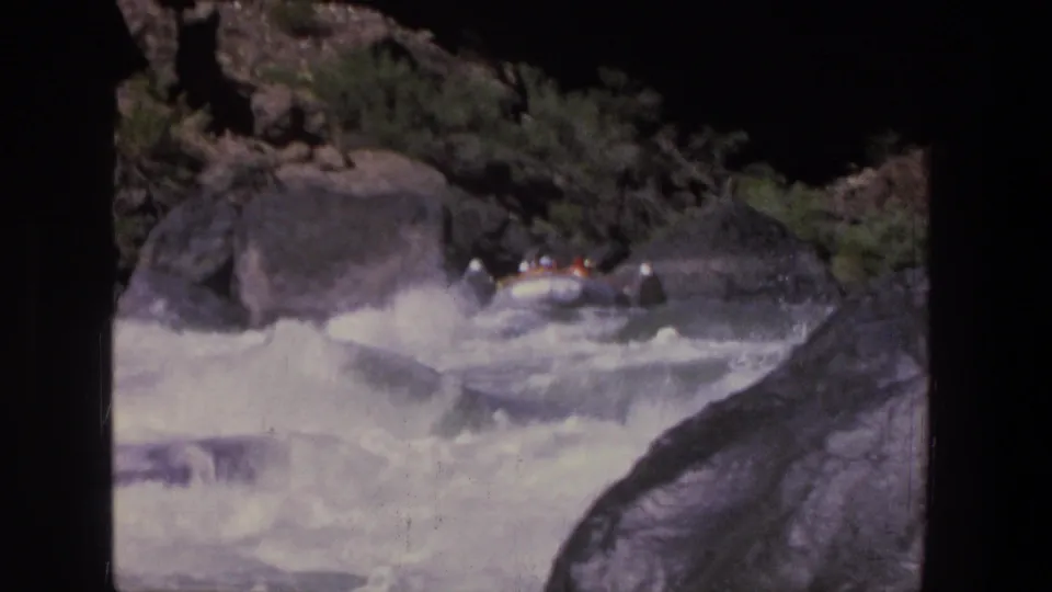 Several people in a small inflatable raft navigate rough Utah whitewater beside large rocks in a 1969 8mm home movie.