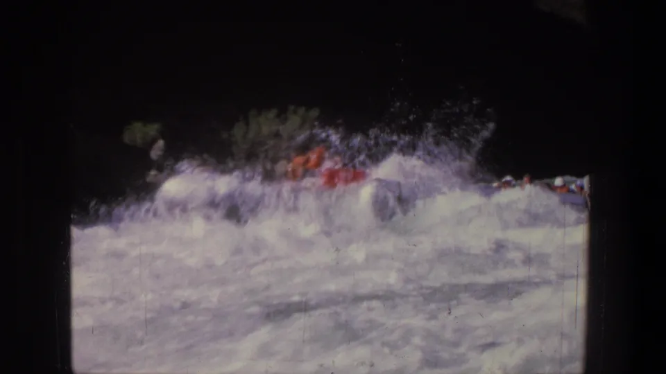 1969 Utah 8mm frame showing a red raft partly obscured by whitewater waves along a shaded riverbank.