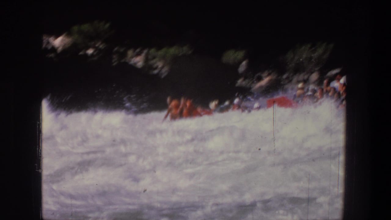 1969 Utah 8mm frame with rafters in a red inflatable raft crossing turbulent whitewater beside a shaded river edge.