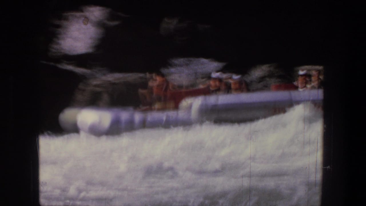 Red inflatable raft with passengers entering white-water rapids near dark rocks.