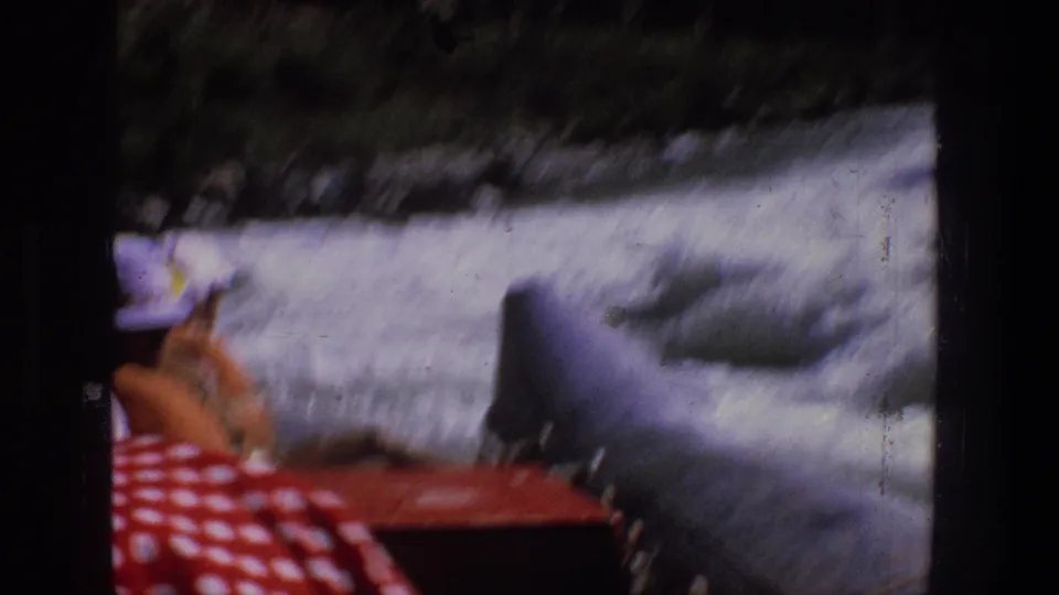 Handheld 1969 Utah raft POV showing a passenger near a raft tube as whitewater rushes past a rocky riverbank.