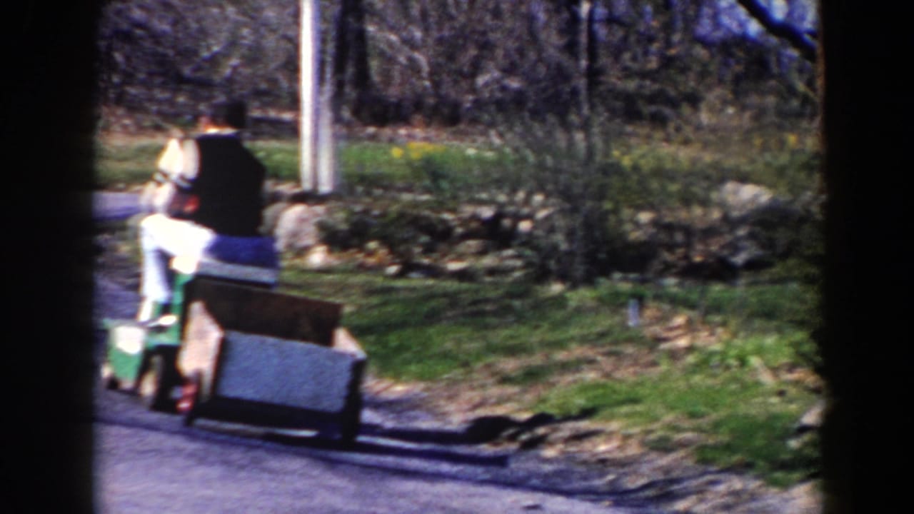 Man drives a green riding mower away down a quiet road while pulling a wooden cart.