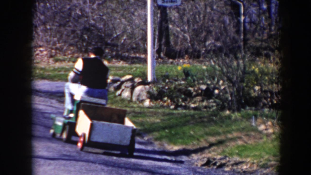The man continues away on the riding mower in the 1964 Pequot Lakes street scene, with a wooden cart trailing behind near stone edging.
