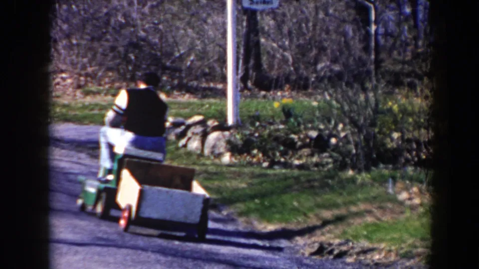 The man continues away on the riding mower in the 1964 Pequot Lakes street scene, with a wooden cart trailing behind near stone edging.