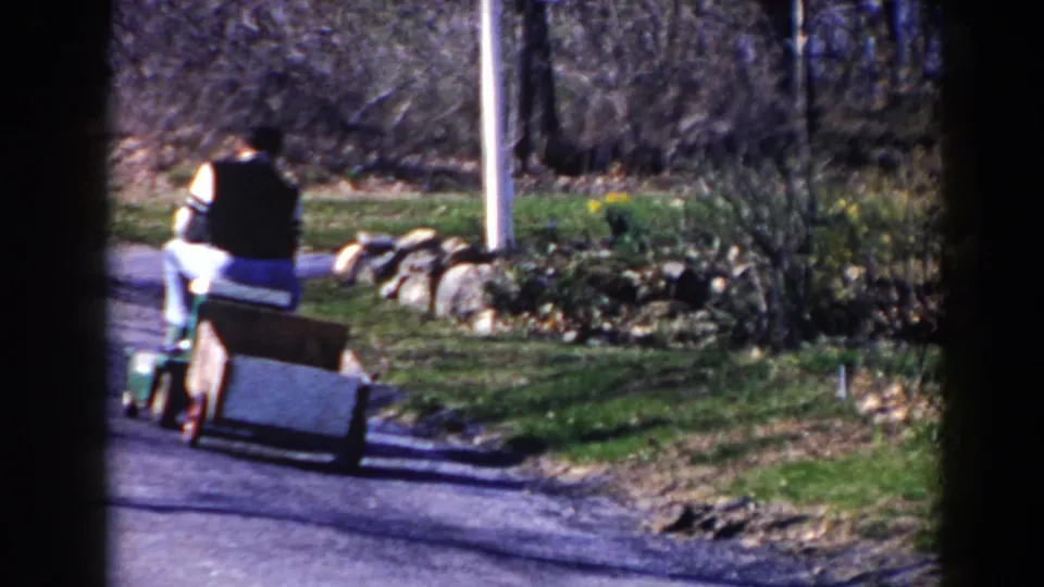 A fixed 8mm view shows the man and tow cart receding along the Pequot Lakes road, with springlike grass and bare trees around the roadside.