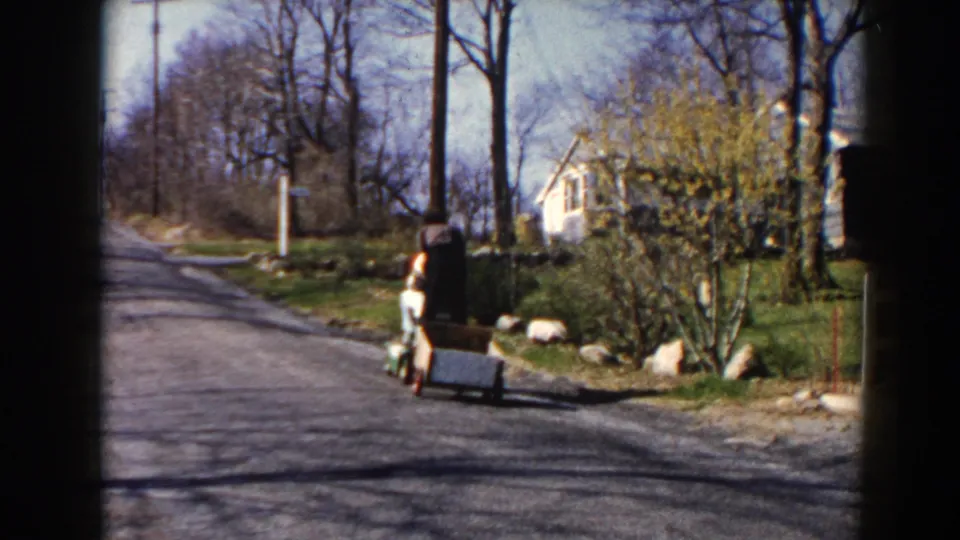 Two women on a small motorized cart pull a low trailer past roadside rocks, shrubs, and houses in 1964 Minnesota footage.