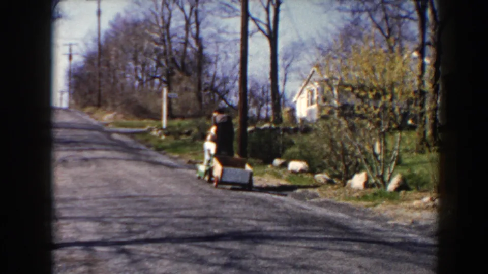 The motorized cart and trailer continue away along a Pequot Lakes road, with houses, trees, and spring greenery in view.