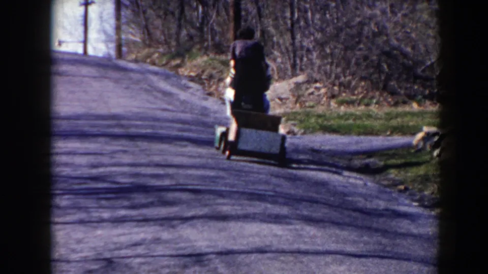 The child continues away along the Pequot Lakes residential road, with the cart, roadside grass, rocks, and bare trees visible.