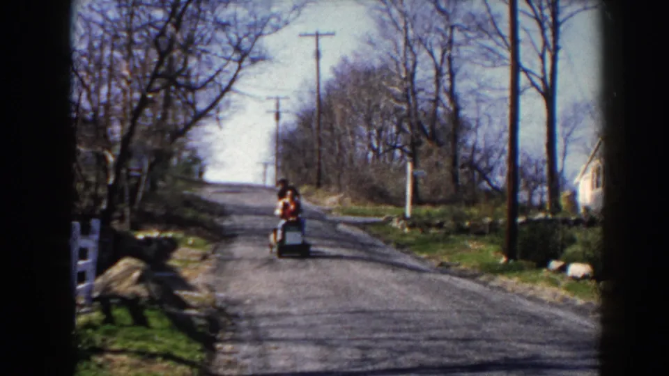 The scooter rider in a red sweater nears the camera on the Pequot Lakes road, with bare trees, poles, and a roadside house edge visible.