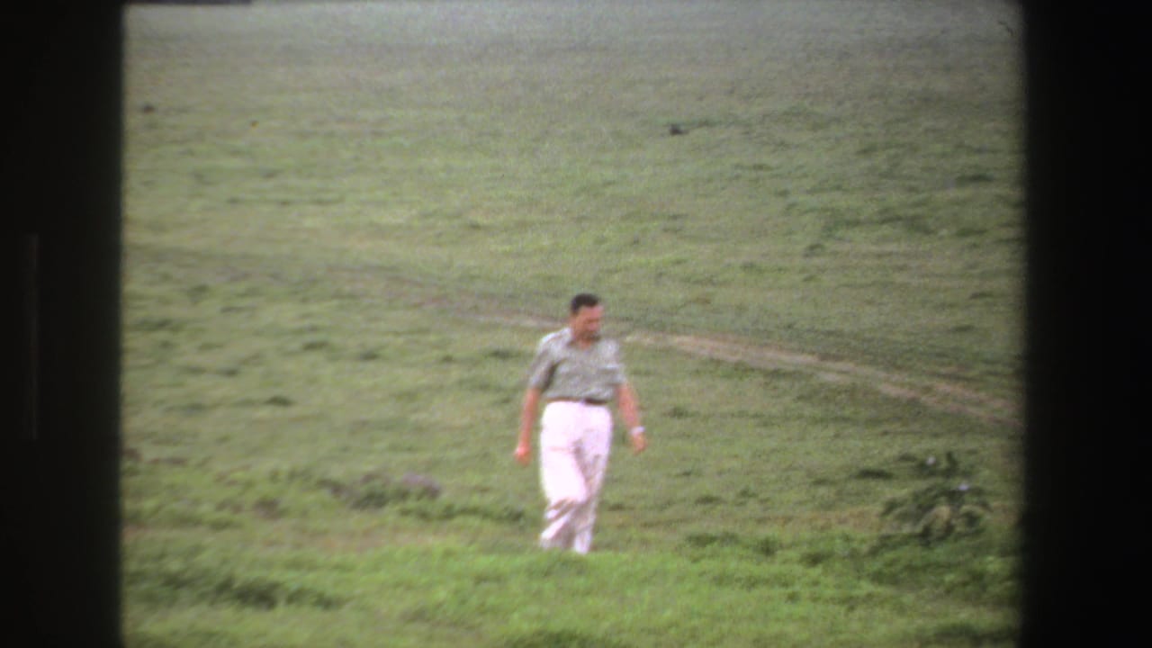 Adult man in light trousers walks across a grassy field with faint tracks behind him.