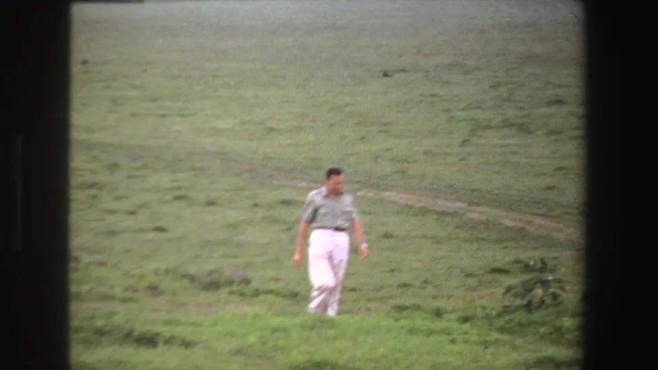 In South Africa home-movie footage from 1969, an adult man in a short-sleeve shirt and white trousers walks through open grassland.