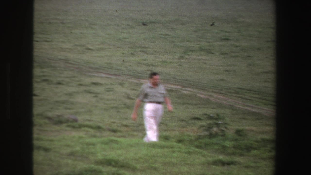 Adult man walks through open grass near a faint dirt path in the background.