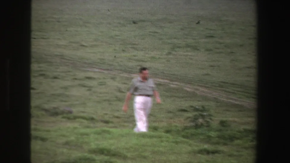 The man continues across the South African grassy field, wearing a short-sleeve shirt and white trousers in soft 8mm color.