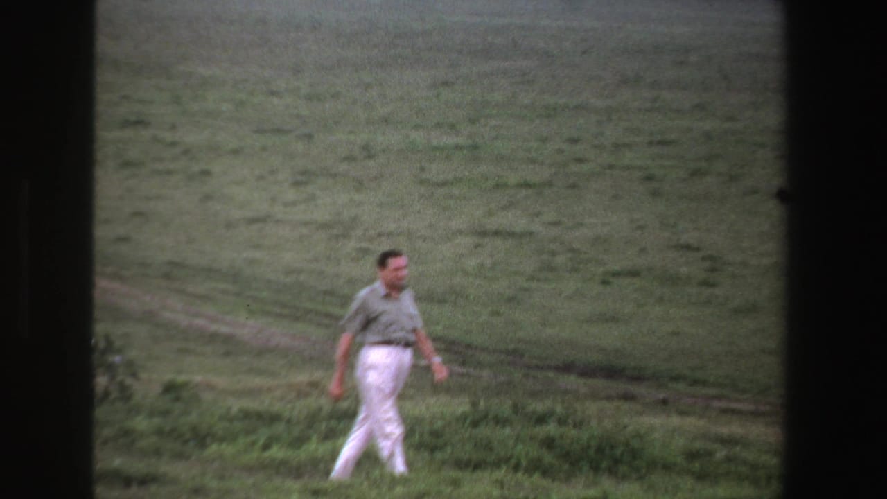 Man in a light shirt and white trousers walking across a grassy field.
