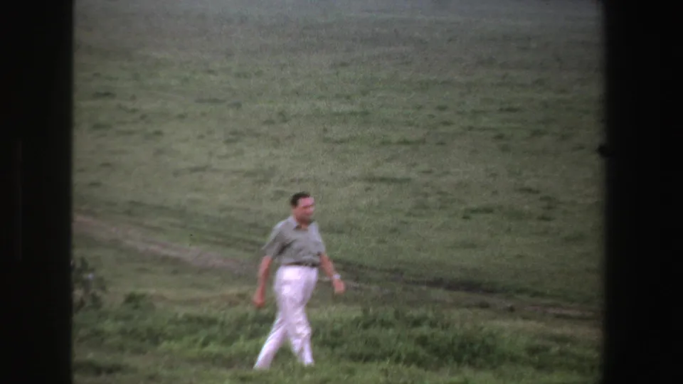 In 1969 South Africa footage, a lone adult man walks across a grassy field with a faint dirt track behind him.