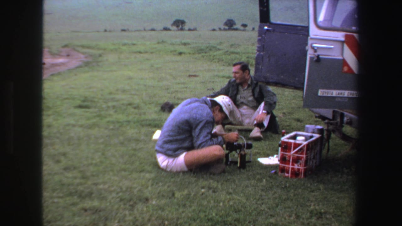 Two men sit near camera equipment beside the rear of a Toyota Land Cruiser in a grassy field.