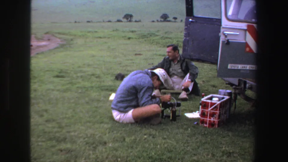 Two adult men pause beside a Toyota Land Cruiser in South Africa, with one working near camera equipment on the grass.